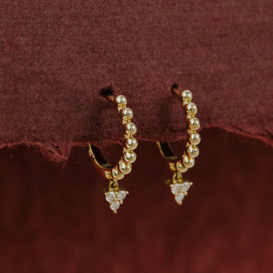 Gold hoop earrings with pearl-like beads and diamond clusters on a burgundy fabric background