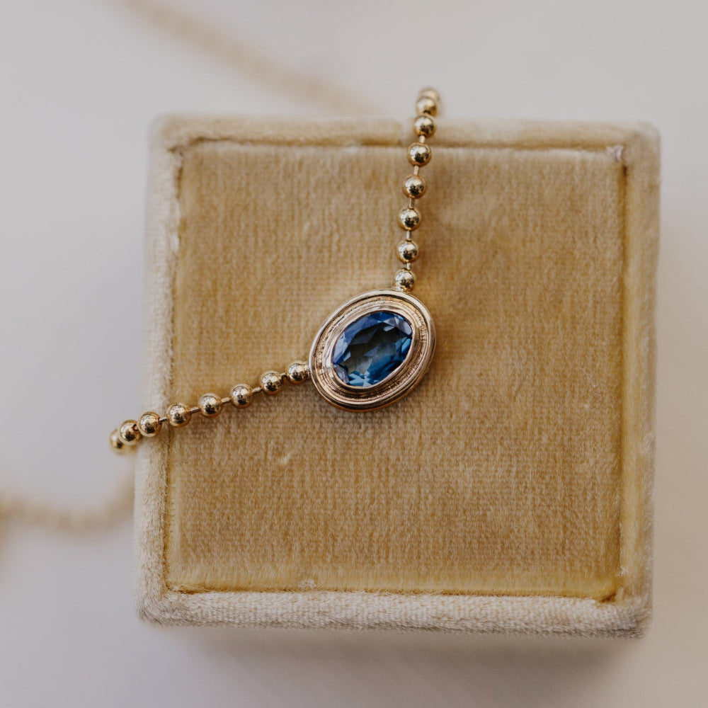 Gold necklace with a blue topaz pendant on a beige velvet cushion