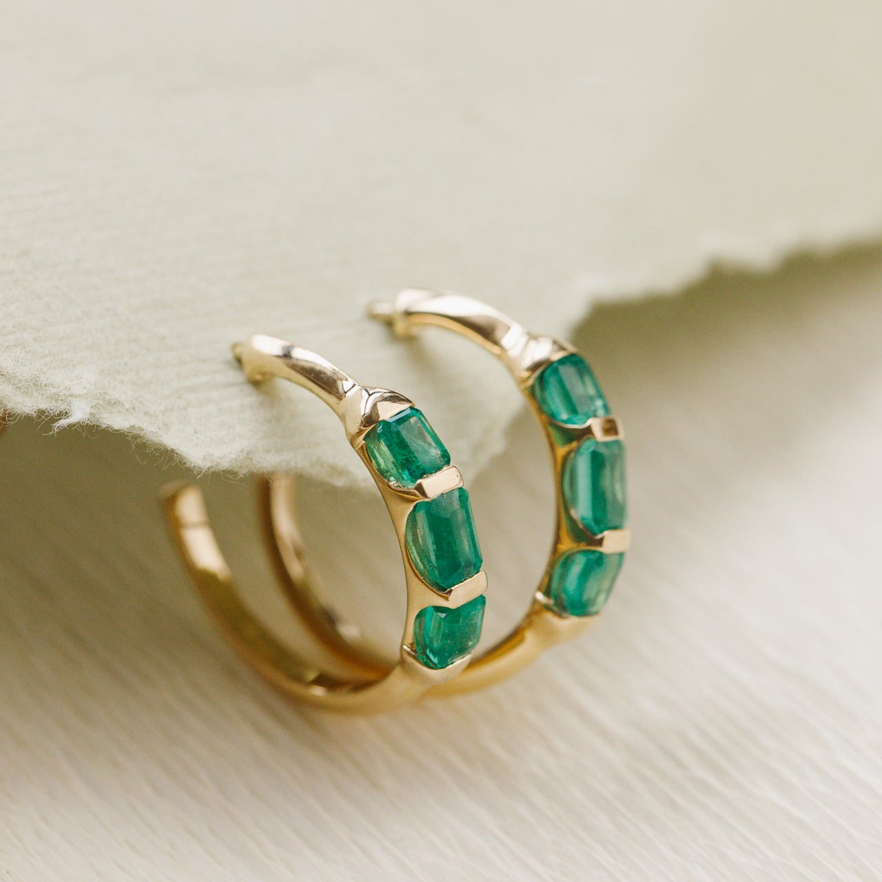 Gold hoop earrings with green emerald gemstones on a light background