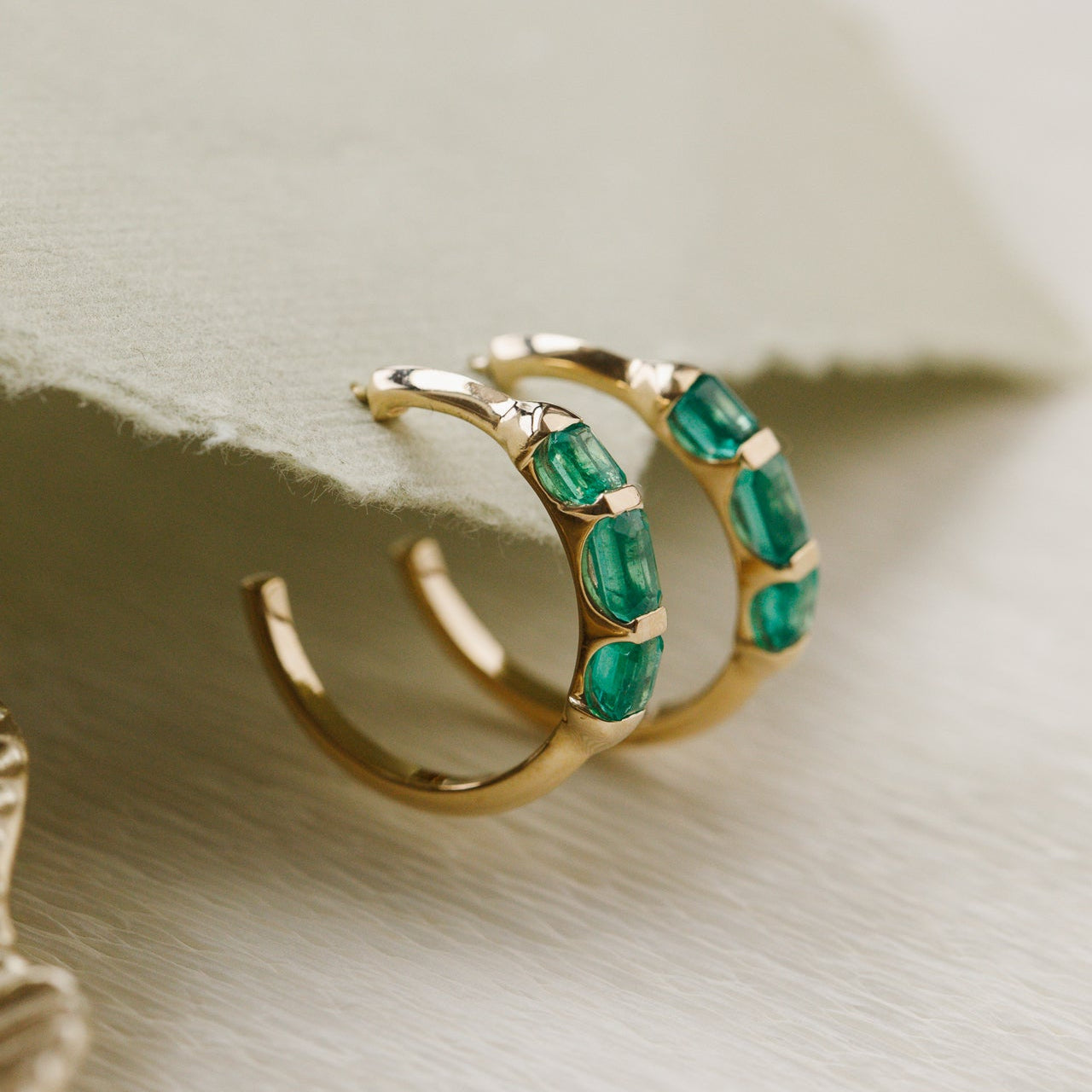 Gold hoop earrings with green emerald gemstones on a beige surface