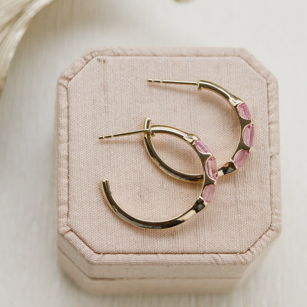 Gold hoop earrings with pink sapphire gemstones on a beige jewelry box