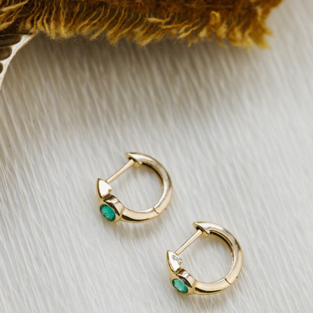 Gold hoop earrings with green emerald gemstones on a textured surface