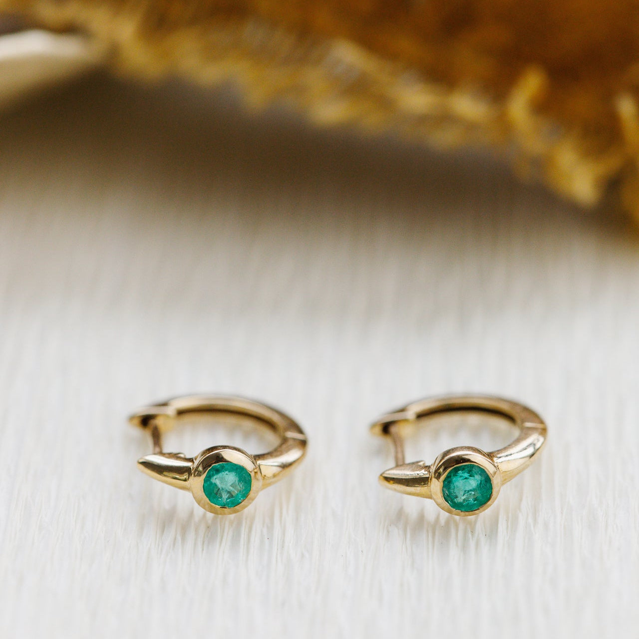 Gold hoop earrings with green emerald gemstones on a textured beige background
