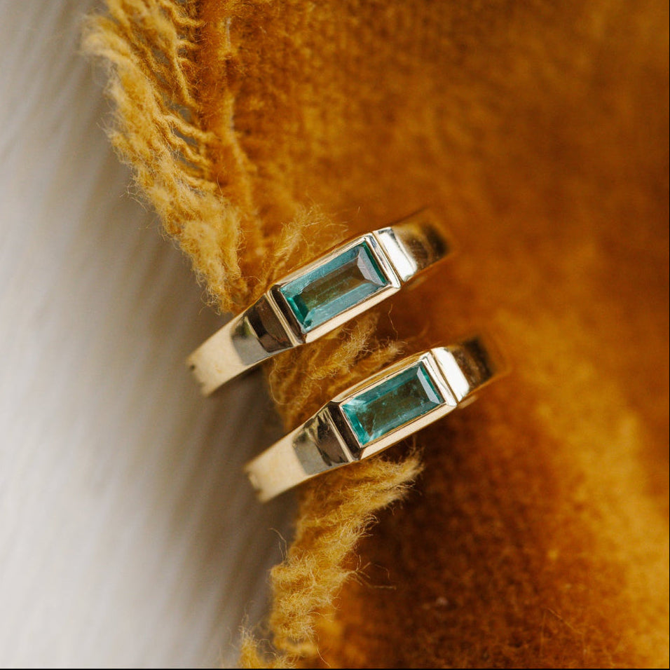 Two gold rings with green emerald gemstones on a textured yellow fabric background