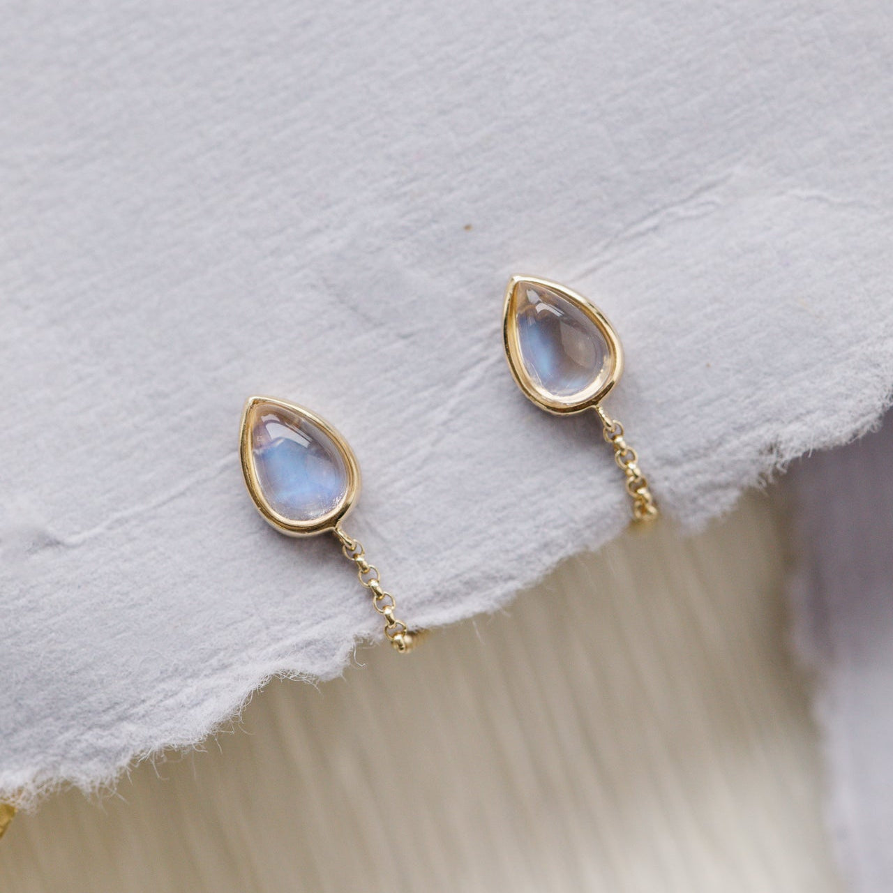 Gold earrings with teardrop-shaped moonstone gemstones on a textured white surface