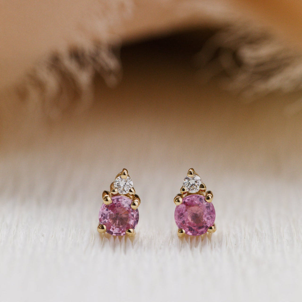 Pair of pink sapphire gemstone earrings with gold and diamond accents on a soft beige background.