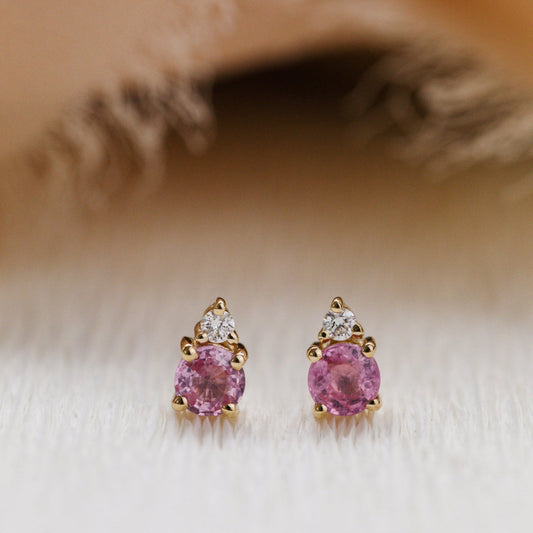 Pair of pink sapphire gemstone earrings with gold and diamond accents on a soft beige background.