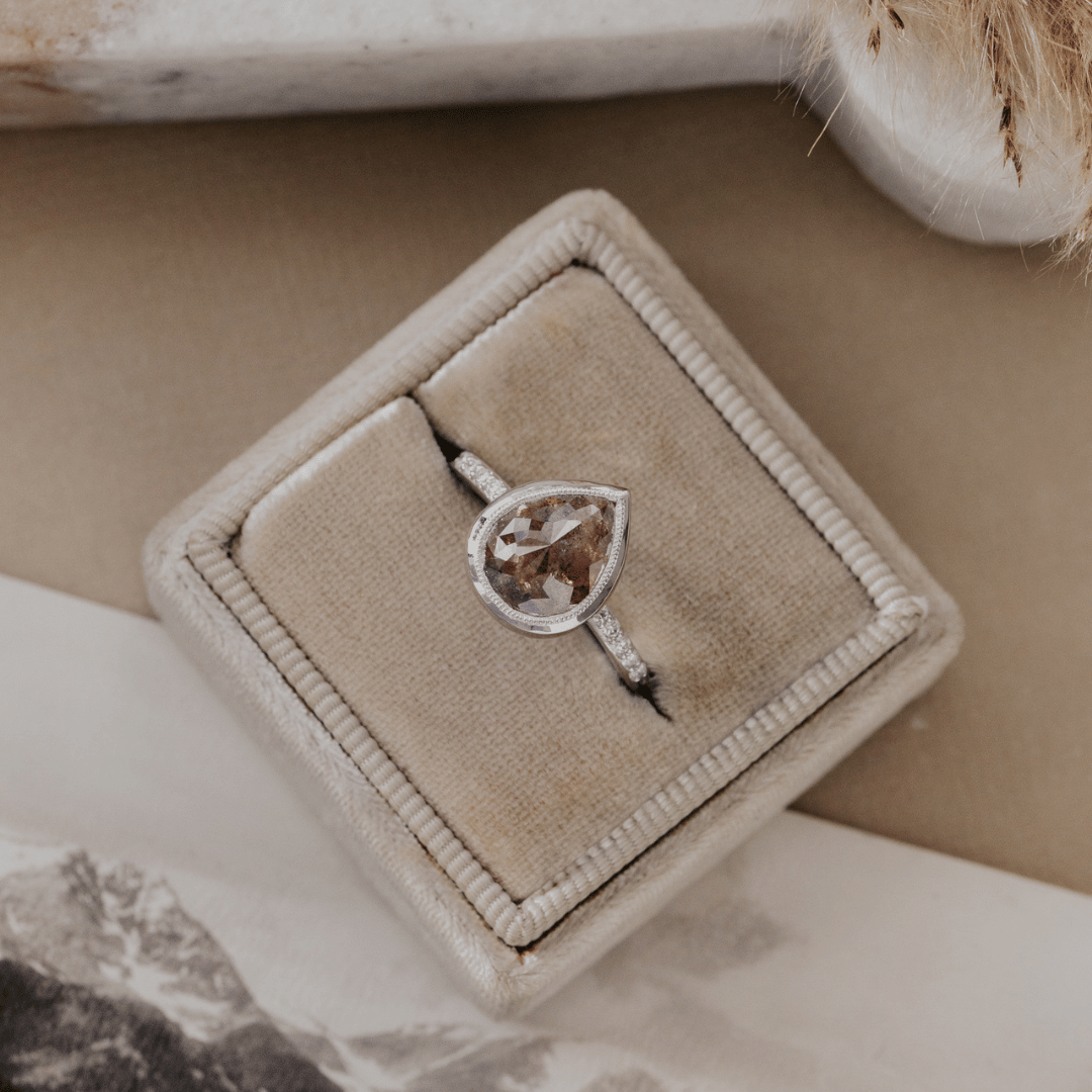 A white gold ring with a pear-shaped brown rustic diamond and small side stones, displayed in a beige velvet ring box on a neutral surface.
