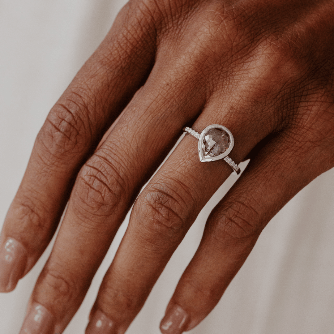 A close-up of a hand wearing a white gold ring with a pear-shaped rustic diamond.