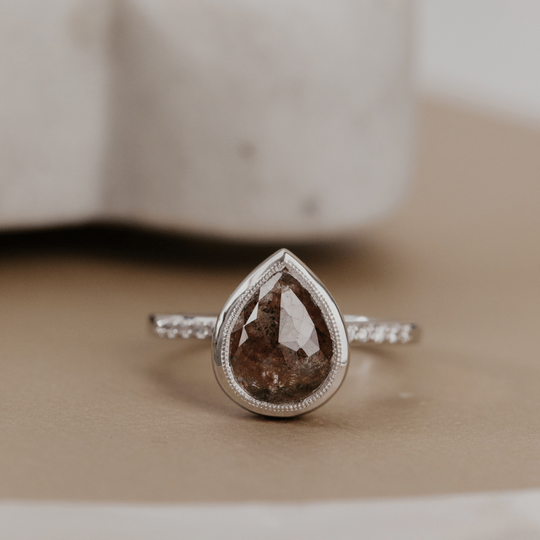 A white gold ring featuring a pear-shaped brown rustic diamond with visible inclusions, set on a textured beige surface with a blurred neutral background.