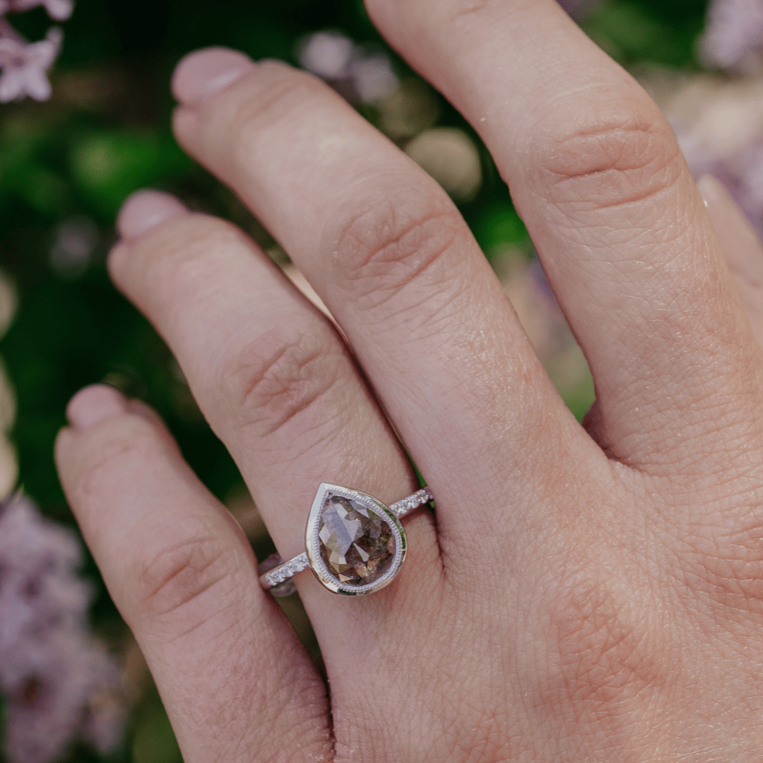 A white gold ring with a pear-shaped smoky brown rustic diamond, worn on a person's ring finger against a soft-focus background of green foliage and pink flowers.