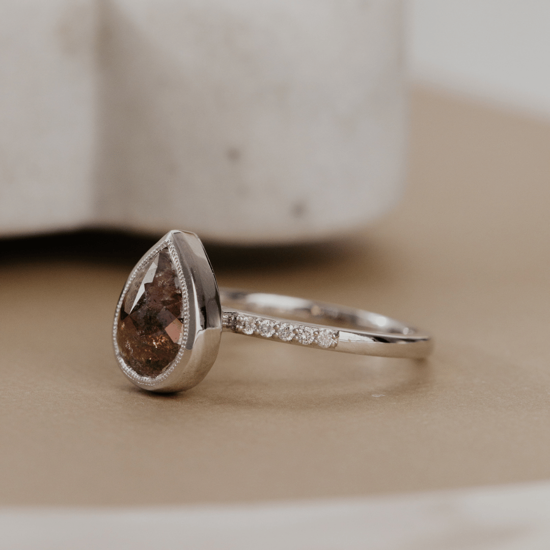 A white gold ring featuring a pear-shaped brown rustic diamond and a micro-pave diamond band, resting on a beige surface with a blurred stone background.