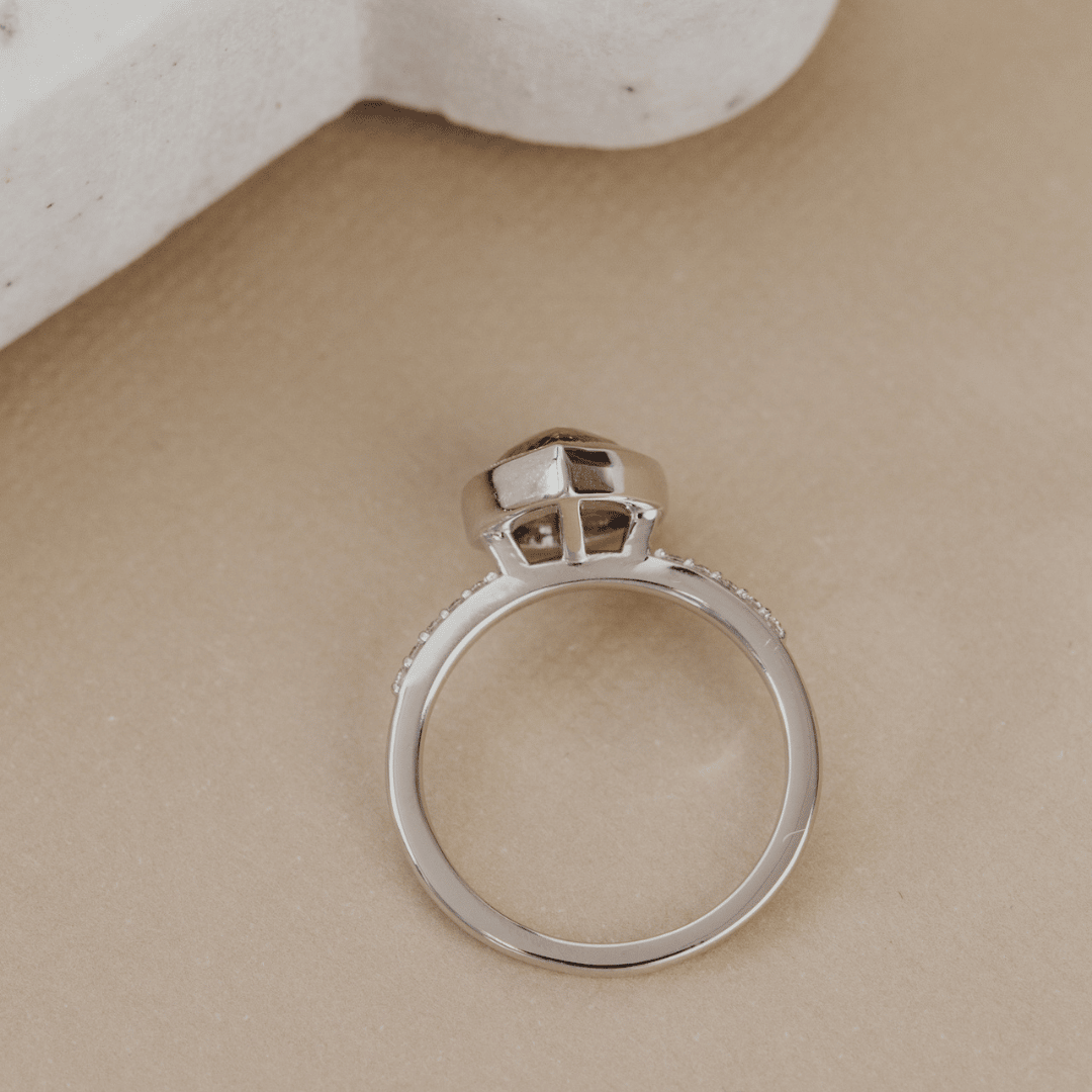 A white gold ring with a geometric crown-like top setting, photographed from above on a warm beige textured surface with a white stone prop in the corner.