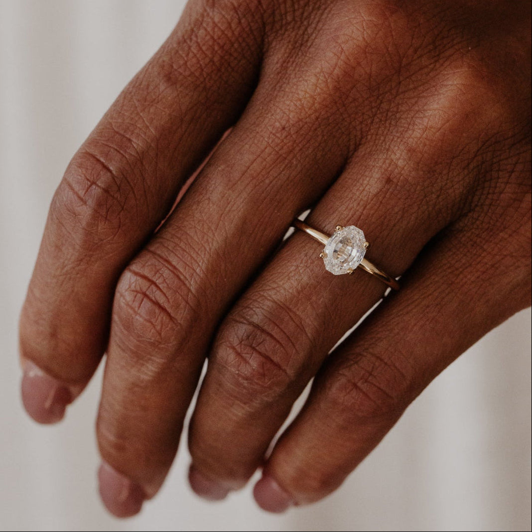 Hand wearing a gold ring with a oval diamond on a neutral background.