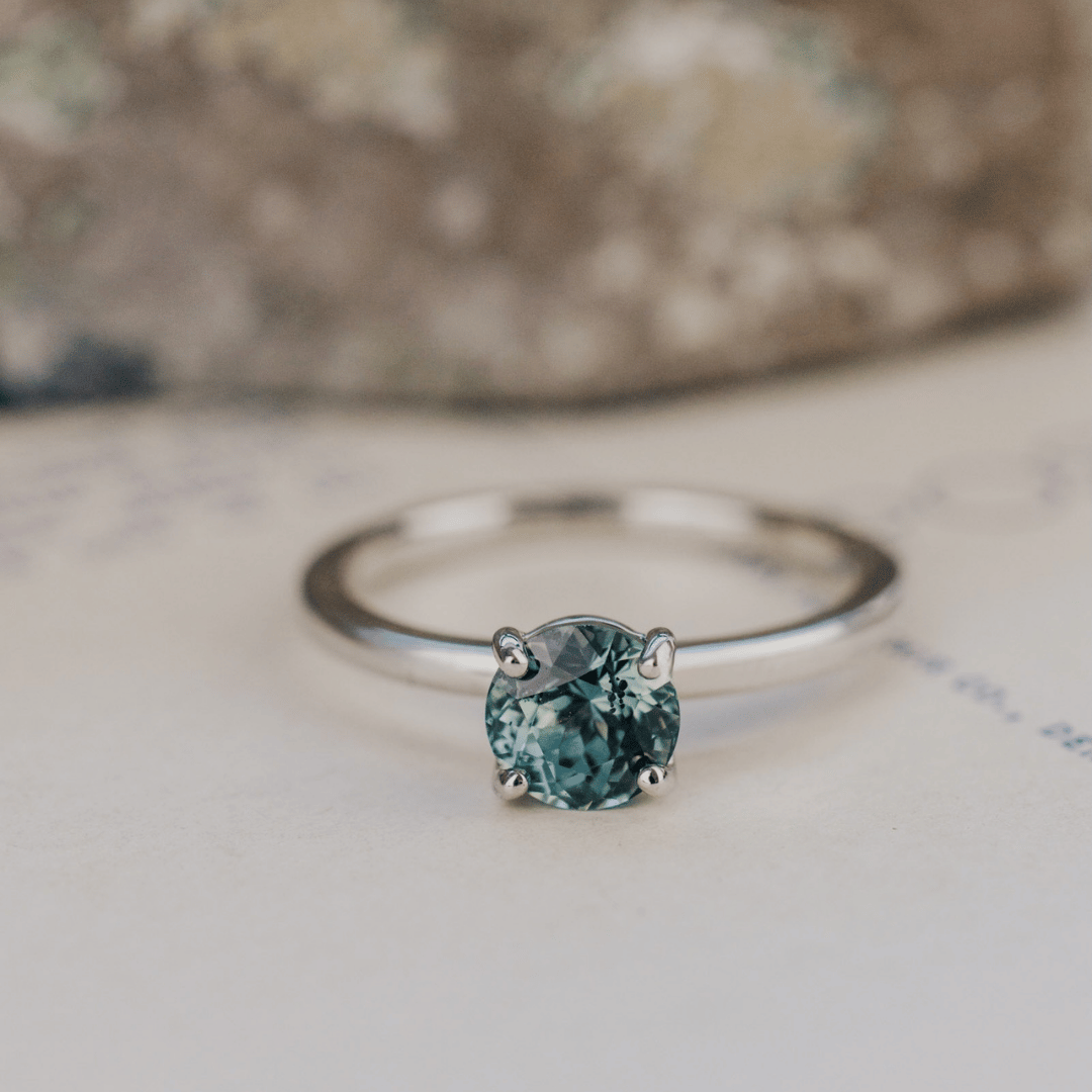 A 14k gold ring featuring a blue sapphire gemstone viewed from straight on, displayed on a textured white surface with a soft-focus background.