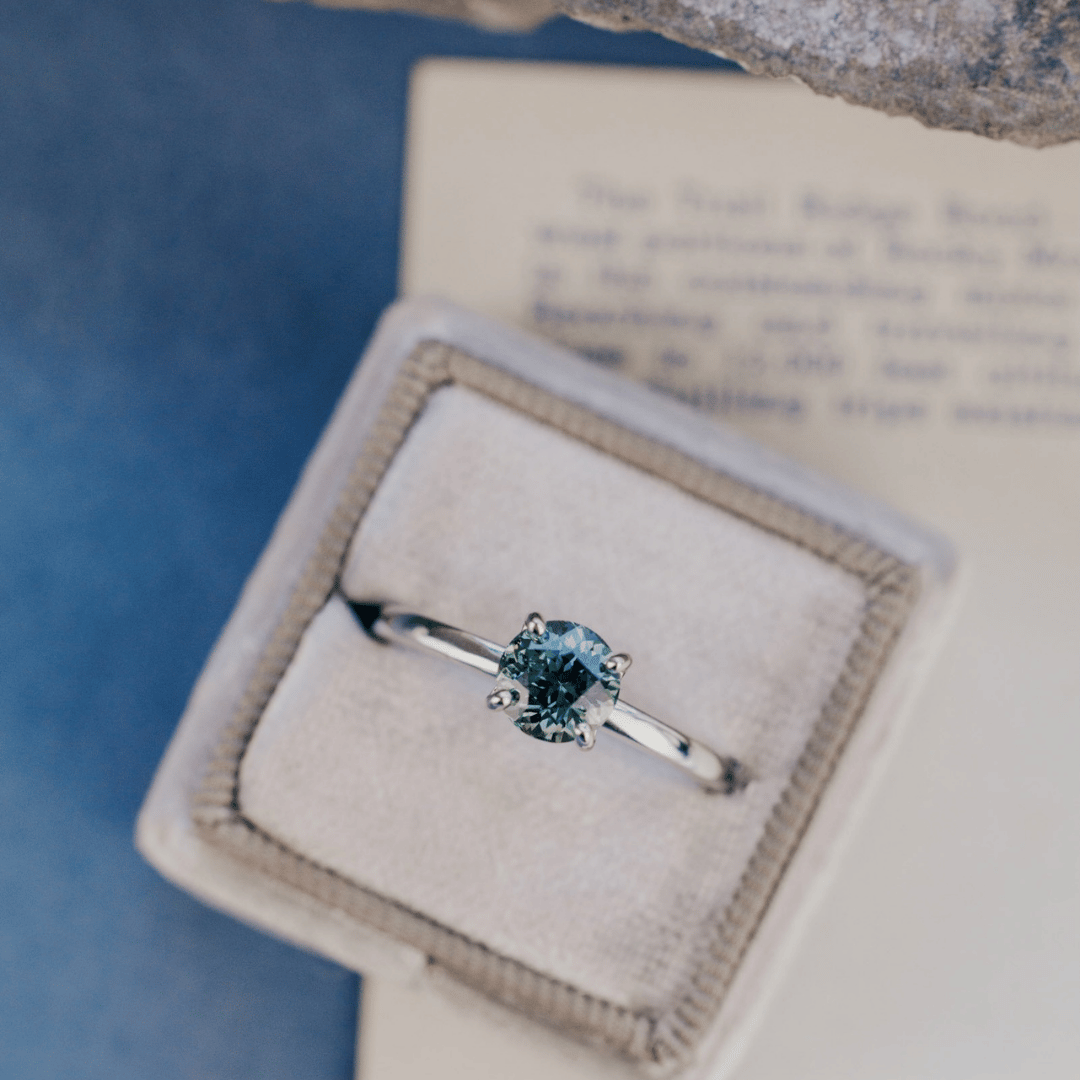 A 14k gold ring featuring a blue sapphire gemstone viewed from a ring box, displayed on a textured white surface with a soft-focus background.