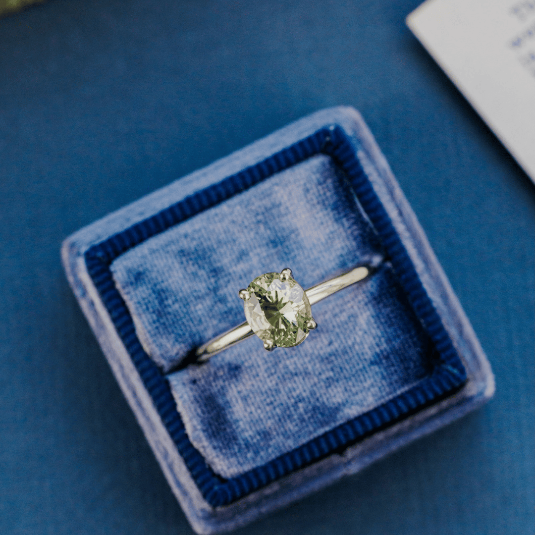 A 14k gold ring featuring a blue/green sapphire gemstone, displayed in a ring box on a textured blue surface.