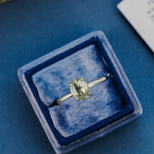 A 14k gold ring featuring a blue/green sapphire gemstone, displayed in a ring box on a textured blue surface.