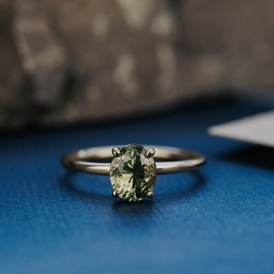 A 14k gold ring featuring a blue/green sapphire gemstone, displayed on a textured blue surface.