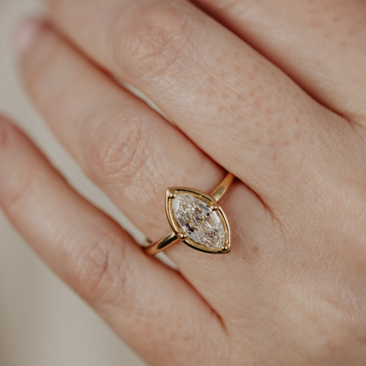Close-up of a hand wearing a gold ring with a marquise lab grown diamond on a neutral background.