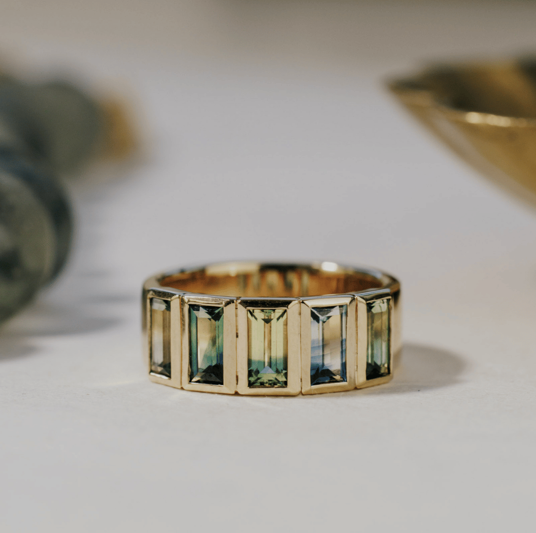 A 14k gold ring featuring multiple blue baguette-shaped sapphire gemstones, displayed on a textured white surface with a soft-focus background.
