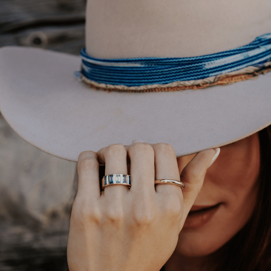 A 14k gold ring featuring multiple blue baguette-shaped sapphire gemstones, displayed on the ring finger of a hand model tipping the brim of her hat with a soft-focus background.