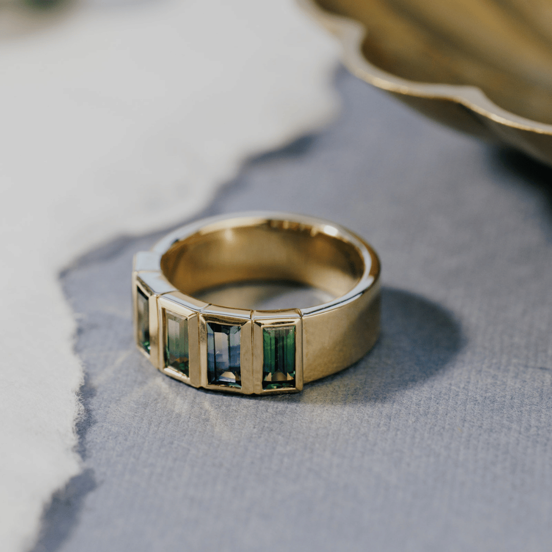 A 14k gold ring featuring multiple blue baguette-shaped sapphire gemstones, displayed on a textured blue surface, angled to the side with a soft-focus background.