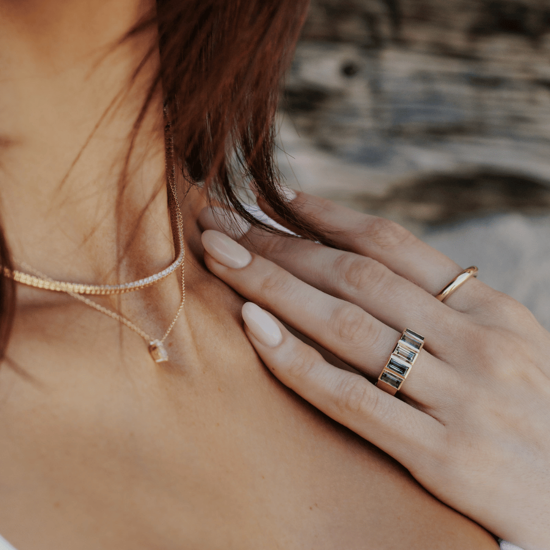 A 14k gold ring featuring multiple blue baguette-shaped sapphire gemstones, displayed on the ring finger of a hand model touching her collar bone.