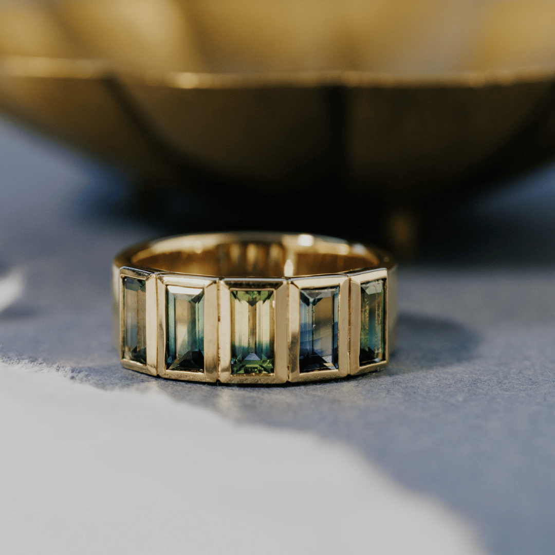 A 14k gold ring featuring multiple blue baguette-shaped sapphire gemstones, displayed on a textured blue and white surface with a soft-focus background.