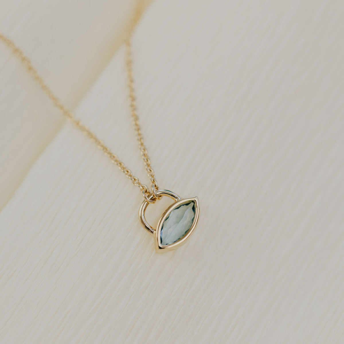 A 14k yellow gold marquis shaped aquamarine lock style necklace on a textured background. 