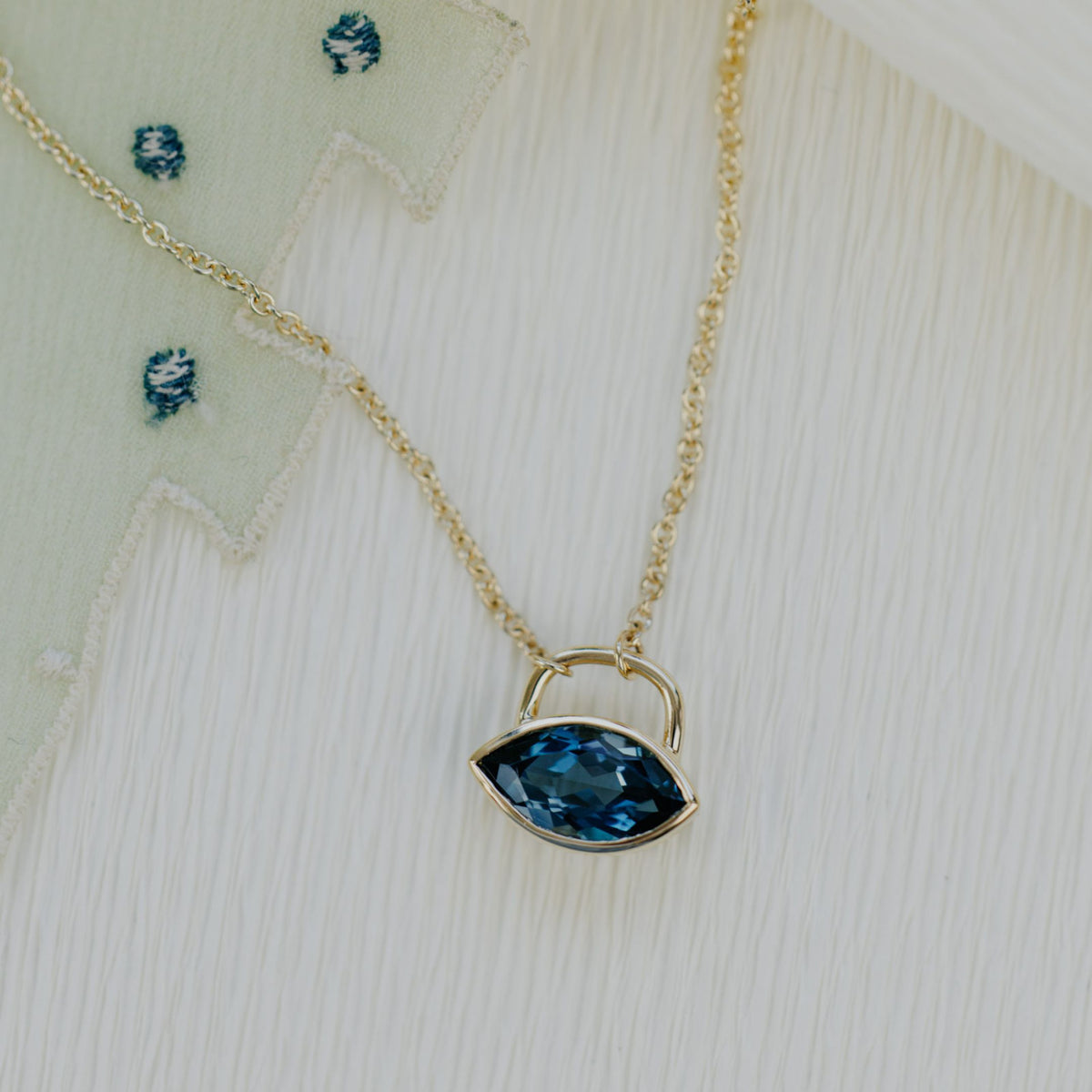 A 14k yellow gold marquise cut blue topaz necklace in a bezel setting on a soft textured background. 