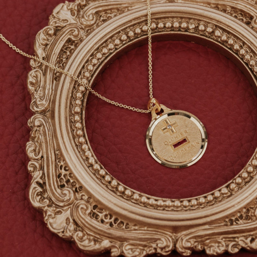 The Medium 18k Yellow Gold Vintage Love Token with Knife Edge and Rounded Bail by A. Augis, a round 18k yellow gold vintage pendant with a knife-edge border, lab grown ruby baguettes, a natural diamond, and the engraved phrase "qu'hier que demain," displayed close-up within an ornate gold oval frame on a deep red surface.