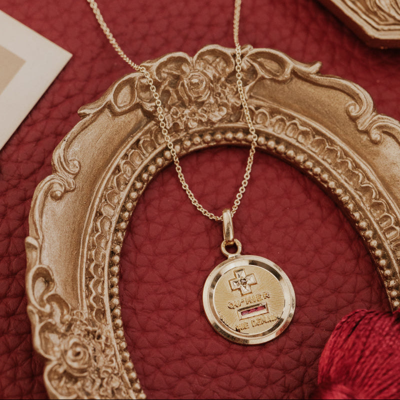 The Medium 18k Yellow Gold Vintage Love Token with Ruby Baguettes by A. Augis, a round 18k yellow gold vintage French love token with lab grown ruby baguettes and engraved text, styled on a gold chain within an ornate gold oval frame on a deep red surface alongside vintage props.