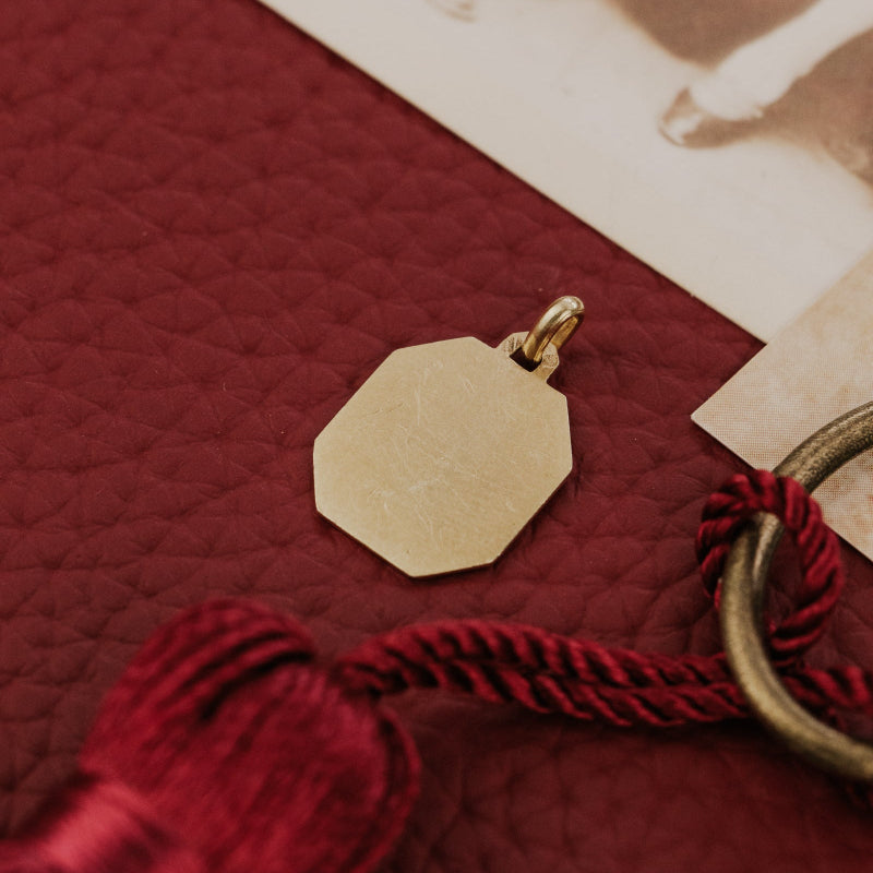 The Medium Rectangular 18k Yellow Gold Vintage Love Token with Rose Border by A. Augis, showing the smooth reverse side of the octagonal 18k yellow gold vintage love token, displayed on a deep red surface alongside a burgundy tassel, vintage key, and sepia photograph.