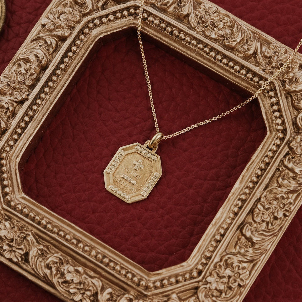 The Medium Rectangular 18k Yellow Gold Vintage Love Token with Rose Border by A. Augis, an octagonal 18k yellow gold vintage French love token with a rose-motif border and engraved text, styled on a gold chain within an ornate gold octagonal frame on a deep red surface.