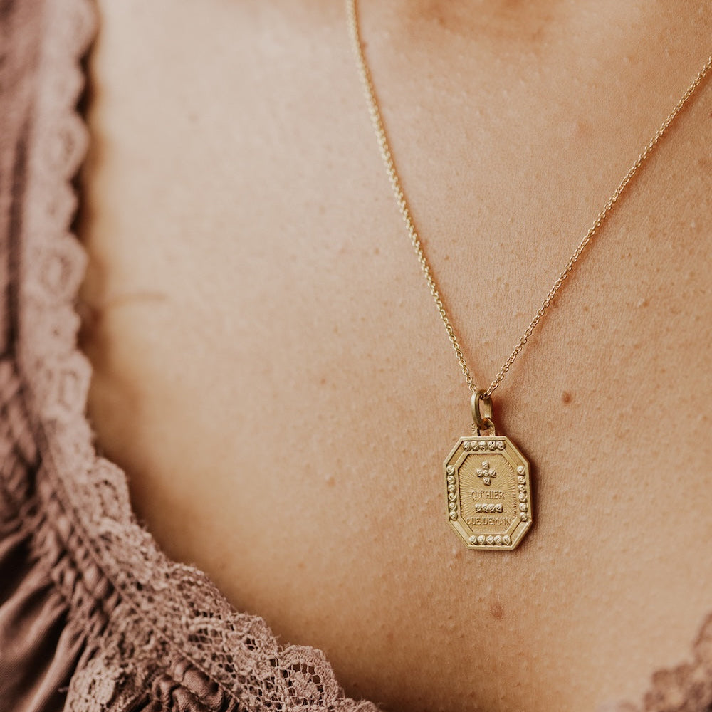The Medium Rectangular 18k Yellow Gold Vintage Love Token with Rose Border by A. Augis, an octagonal 18k yellow gold vintage pendant with a rose-motif border and the engraved phrase "qu'hier que demain," shown at an angle on a gold chain against a model's décolletage.