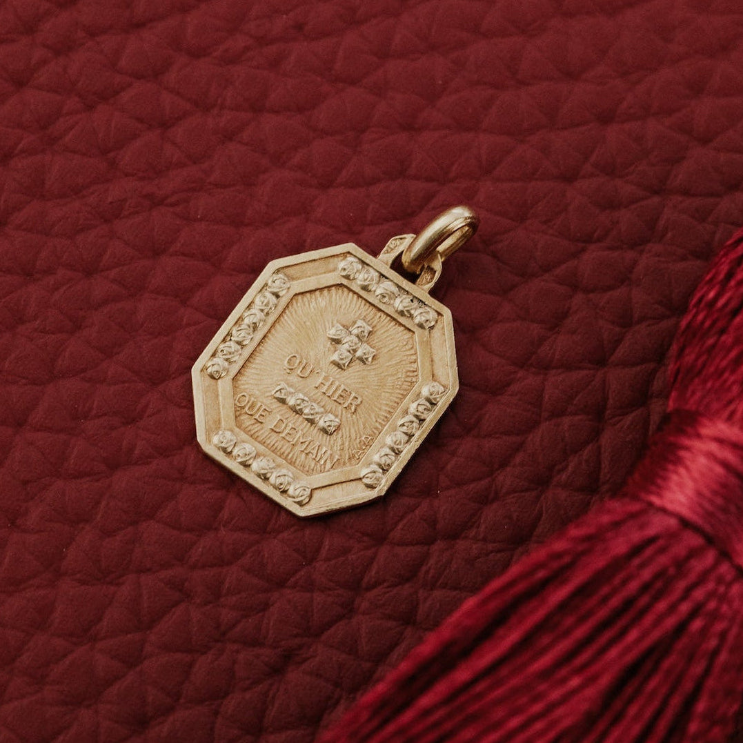The Medium Rectangular 18k Yellow Gold Vintage Love Token with Rose Border by A. Augis, an octagonal 18k yellow gold vintage French love token with a rose-motif border and the engraved phrase "qu'hier que demain," displayed on a deep red surface alongside a burgundy tassel.