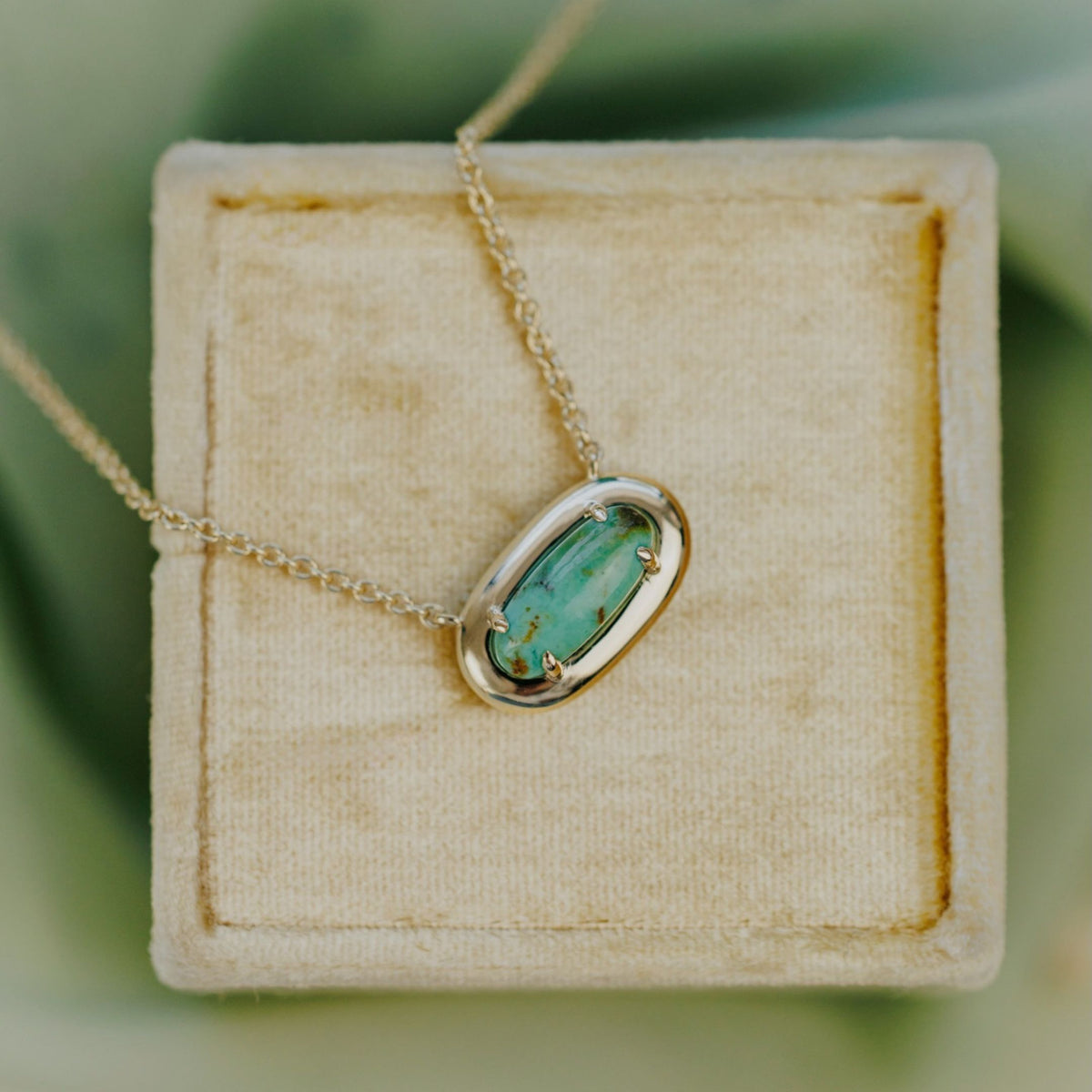 A 14k yellow gold, mini elongated oval turquoise necklace in a metal border setting set on a velvet jewelry box. 