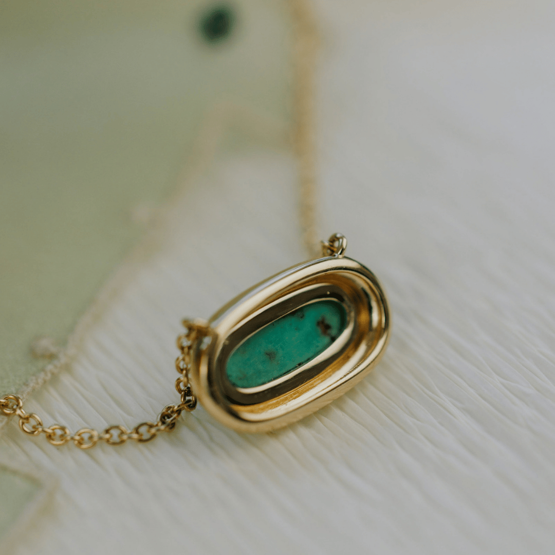 The backside of a 14k yellow gold, mini elongated oval turquoise necklace in a metal border setting set a textured background. 
