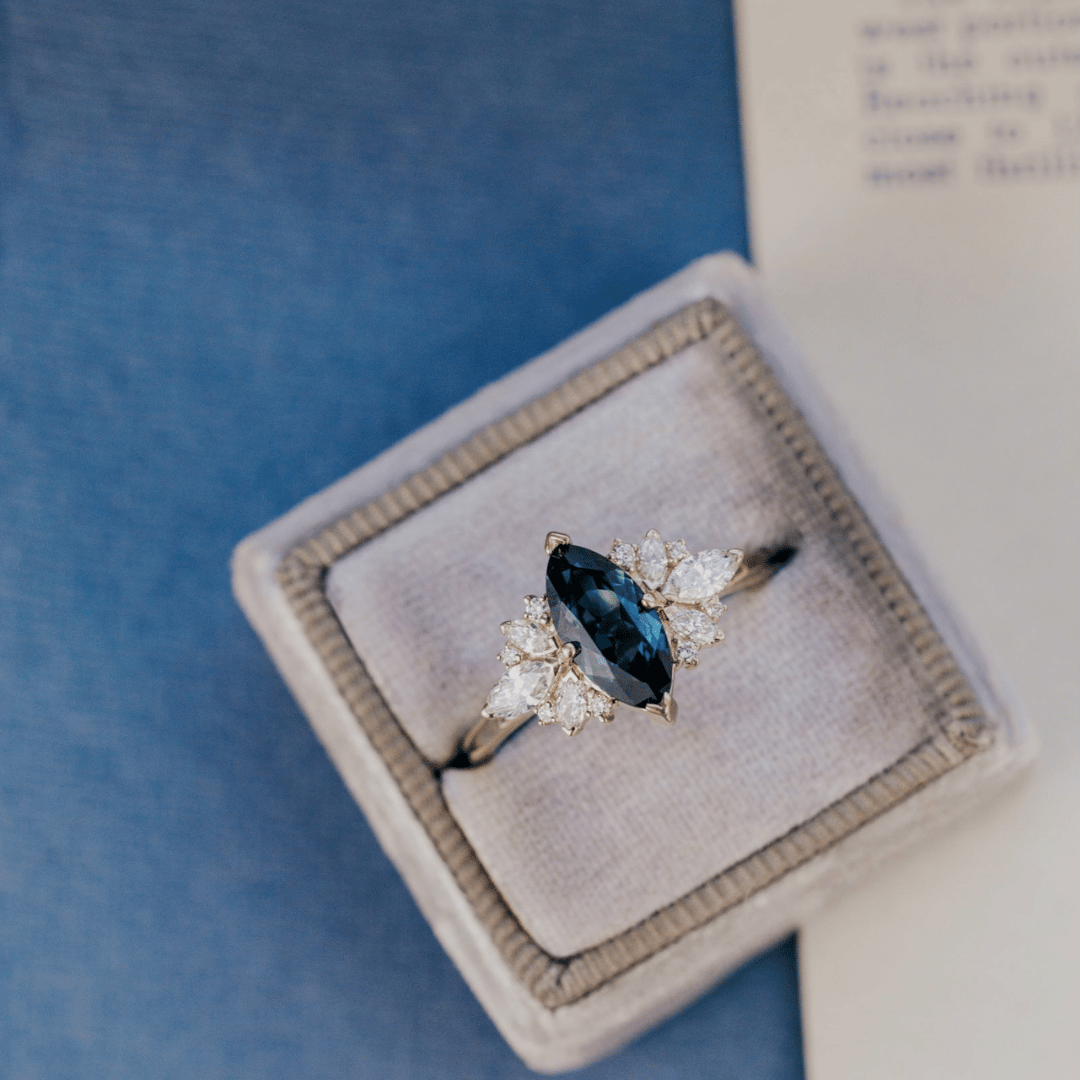 A 14k gold ring featuring a blue sapphire gemstone viewed a ring box, displayed on a textured blue and white surface.