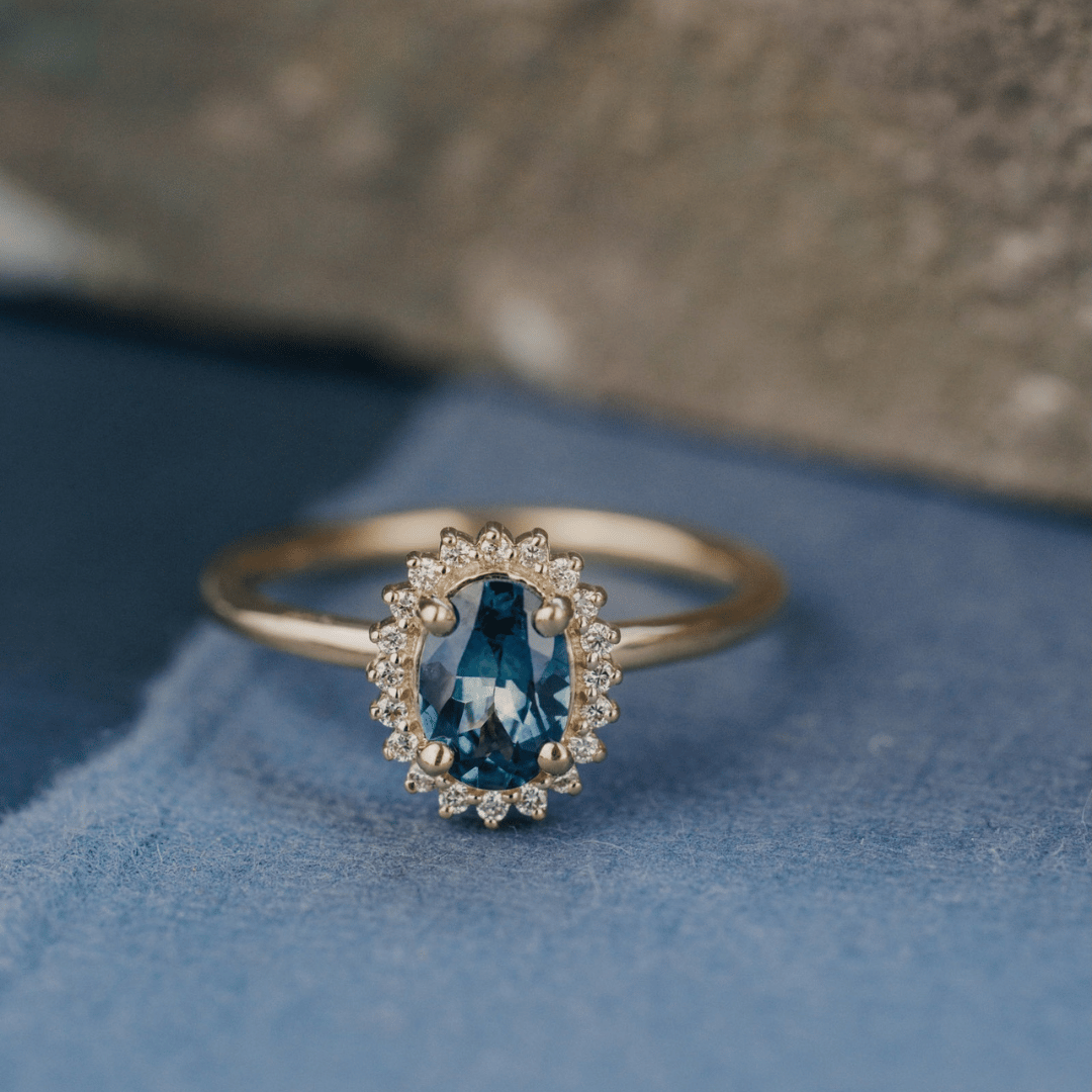 A 14k gold ring featuring a blue sapphire gemstone viewed from straight on, displayed on a textured blue surface with a soft-focus background.