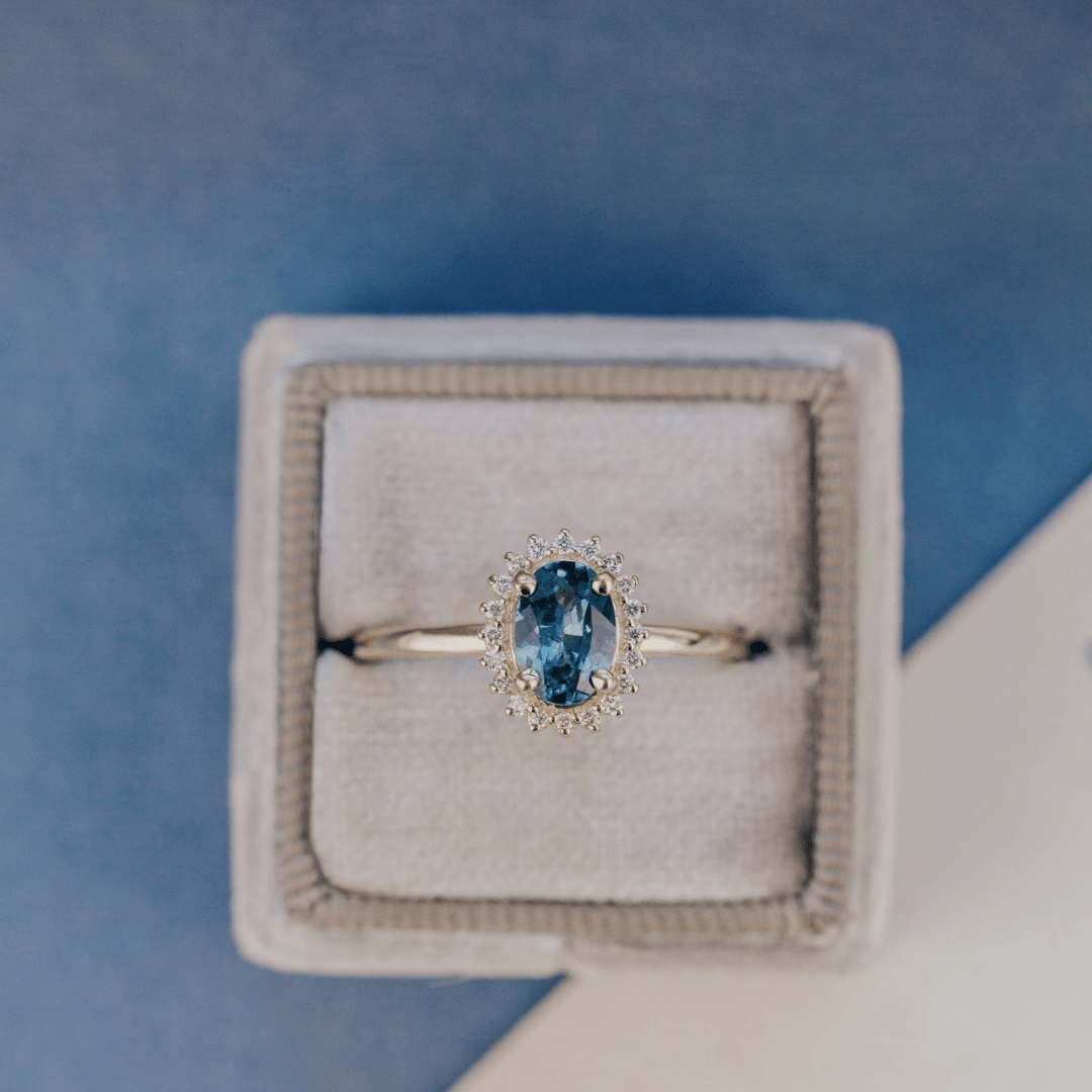 A 14k gold ring featuring a blue sapphire gemstone viewed in a ring box, displayed on a textured blue and white surface.