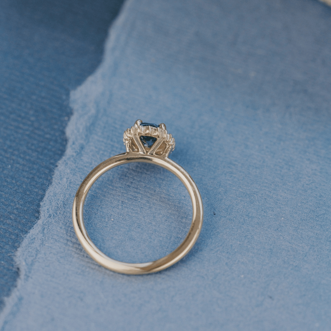 A 14k gold ring featuring a blue sapphire gemstone viewed from the top, displayed on a textured blue surface.