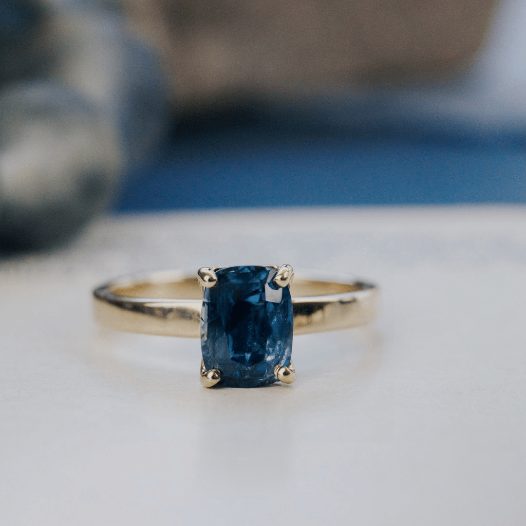 A 14k gold ring featuring a dark blue sapphire gemstone, displayed on a white surface with a soft-focus background.