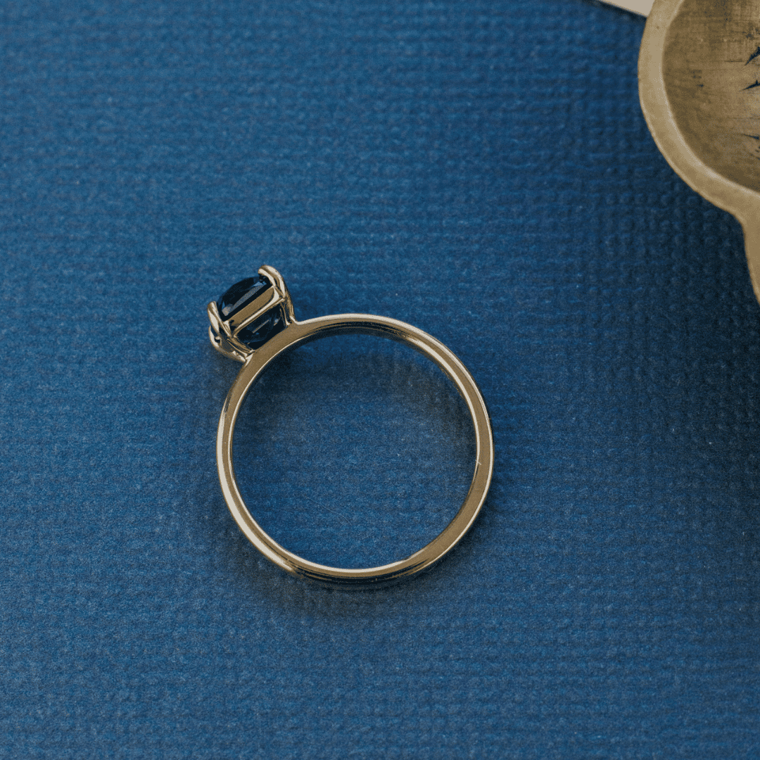A 14k gold ring featuring a dark blue sapphire gemstone from a top view, displayed on a textured blue surface.