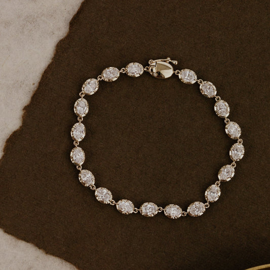 Gold tennis bracelet with oval lab grown diamonds on a textured brown surface.