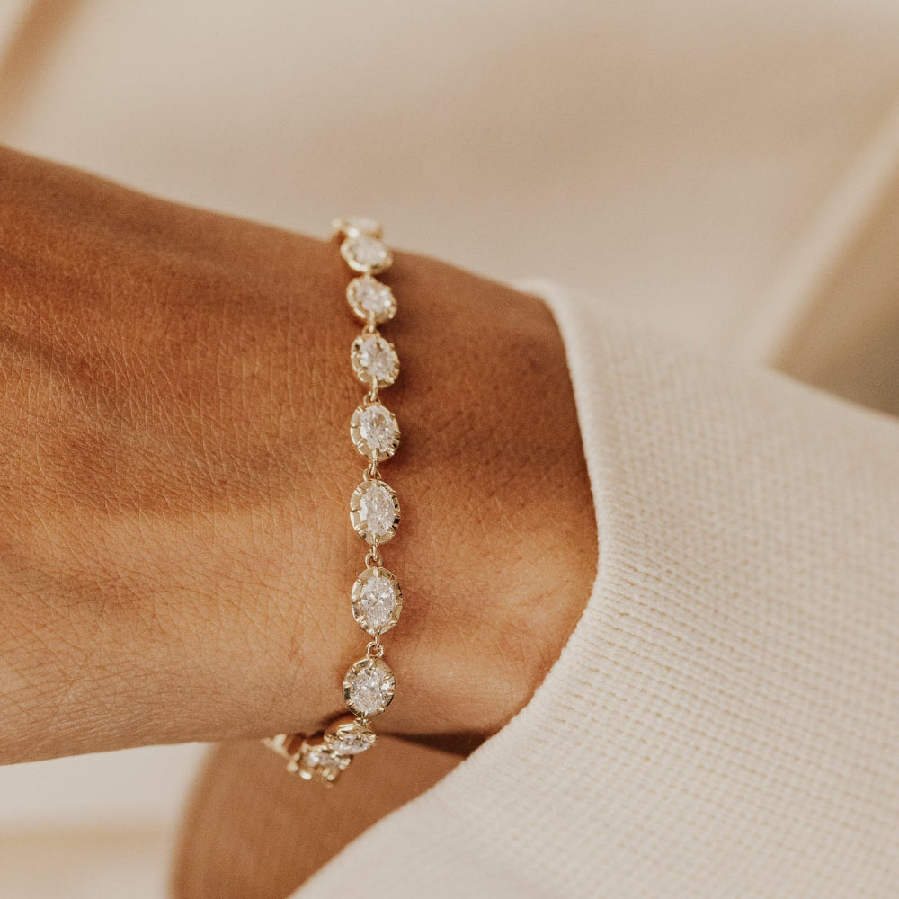 Gold tennis bracelet with oval lab grown diamonds on a wrist against a beige background.