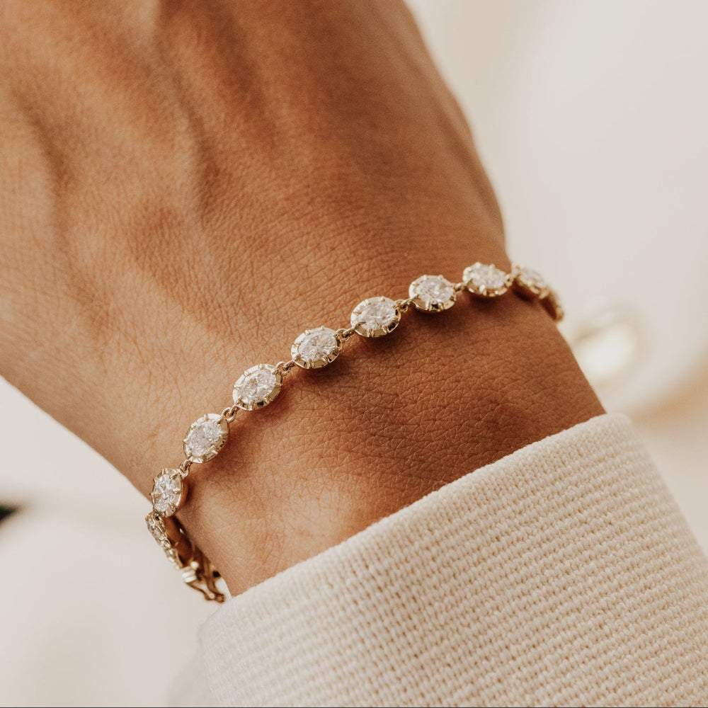 Gold tennis bracelet with oval lab grown diamonds on a wrist against a neutral background.