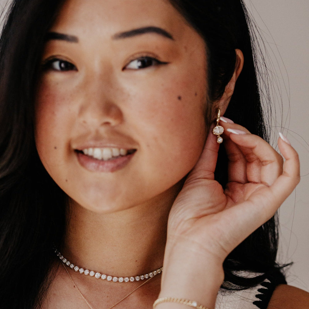 Woman adjusting a gold huggieearring with lab grown diamond dangles on a neutral background.