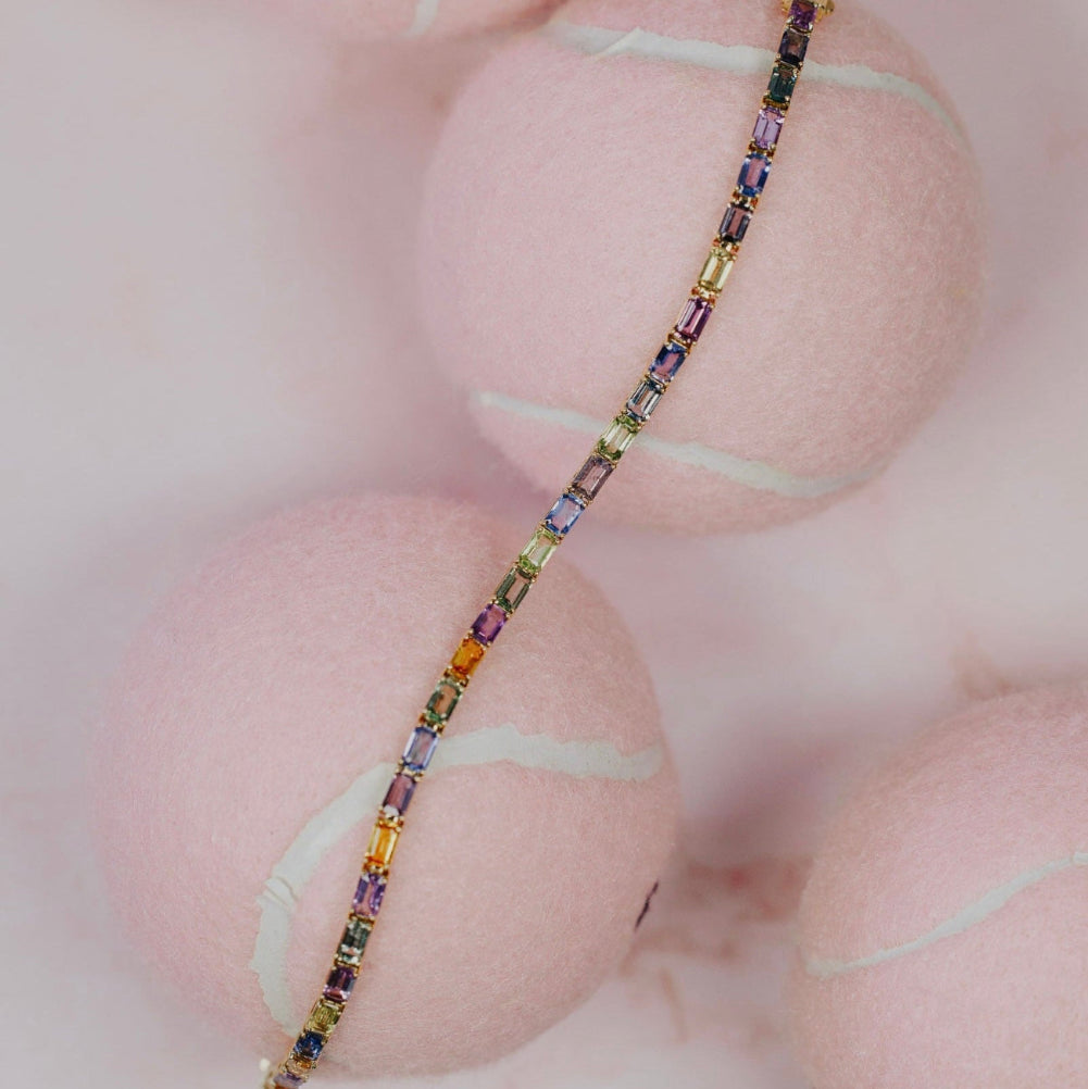The Pastel Emerald Cut Sapphire Tennis Bracelet in 14k yellow gold, featuring 8.75 cttw emerald cut natural sapphires in a variety of pastel colors secured in prong settings, shown unclasped across pink tennis balls.
